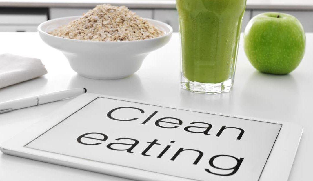 a tablet with the text clean eating written in it and a bowl with oatmeal cereal, a glass with a green smoothie and an apple on the kitchen table